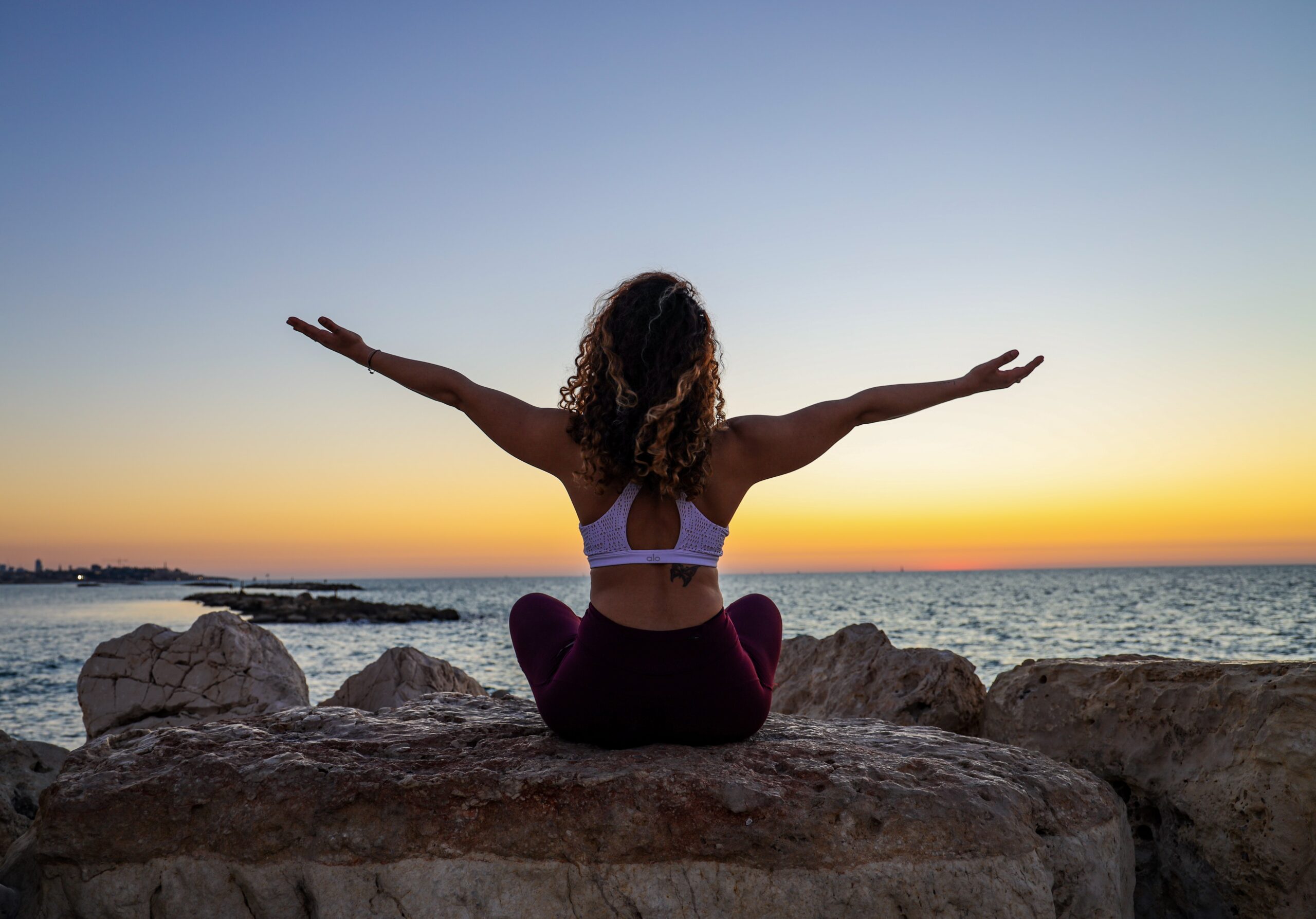 woman sitting on a rock with arms outstretched, facing a sunset