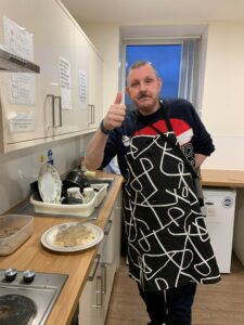 male resident wearing black apron with white pattern, standing with thumbs up in the kitchen next to plate of mushroom stroganoff