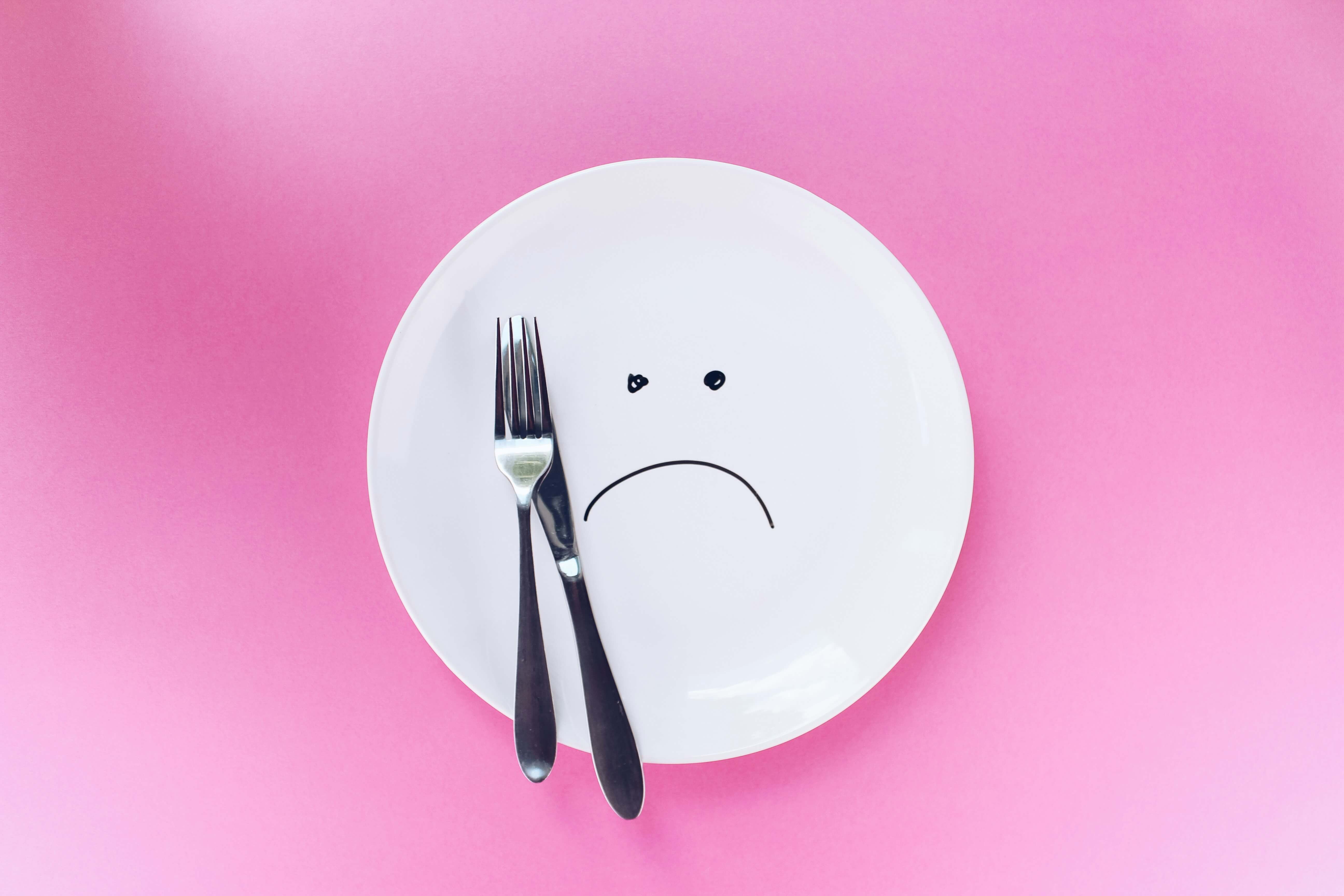 White plate with a sad face drawn on next to a knife and fork on a pink background, representing the eating disorder ARFID (Avoidant/Restrictive Food Intake Disorder).