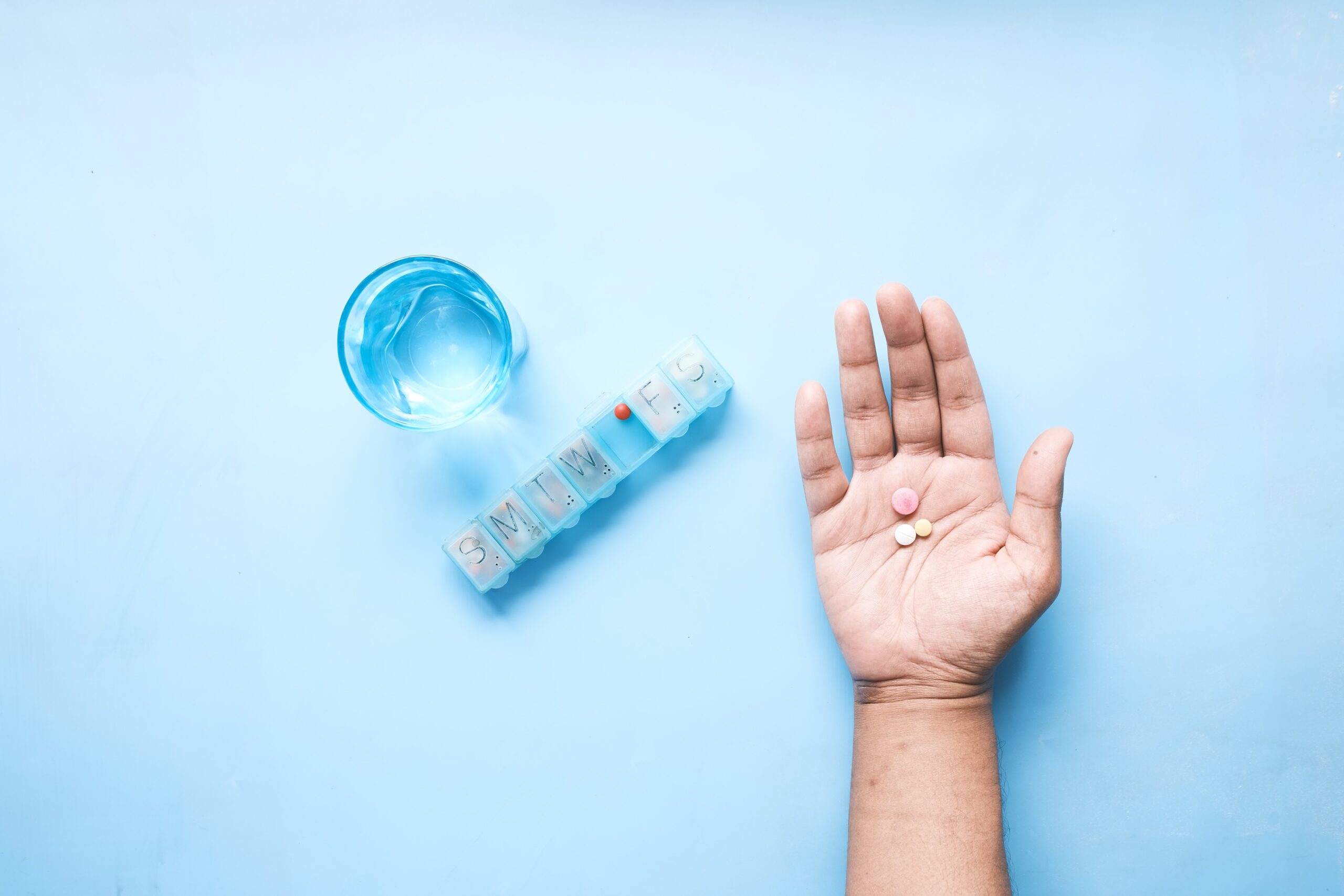 glass of water, medication box and open palm with medication on light blue background