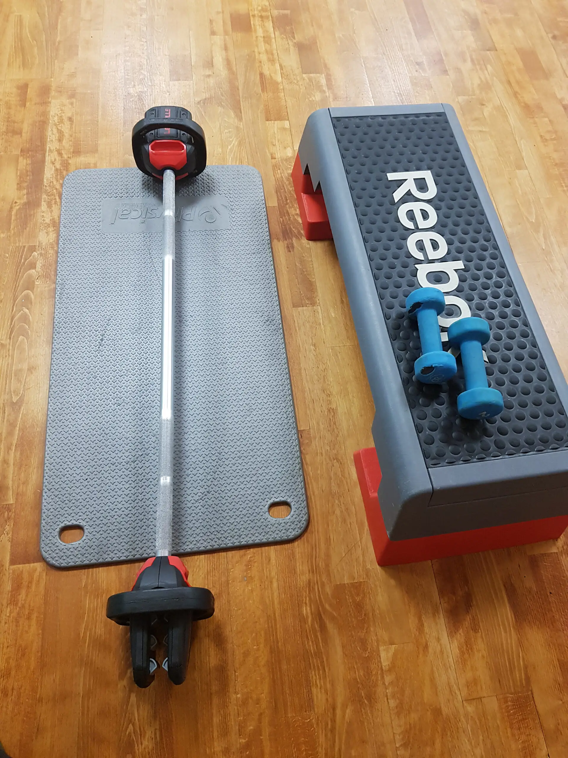 reebok step with blue dumbells next to black exercise mat with weighted barbell laid on top