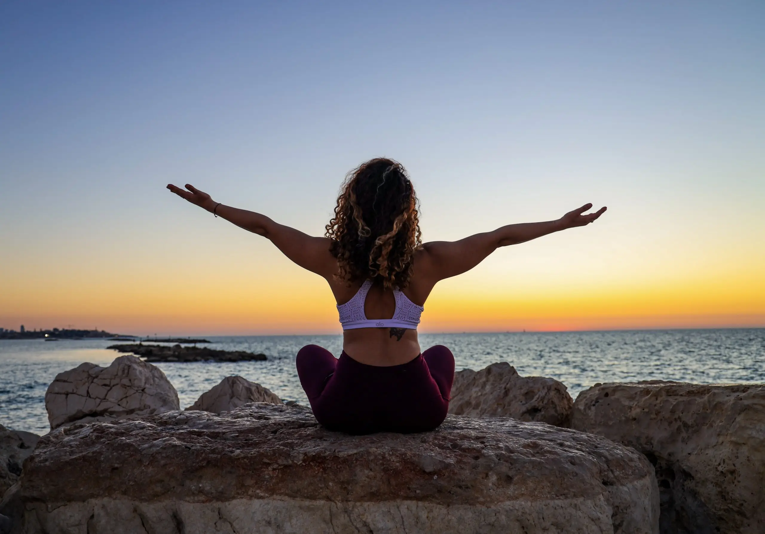 woman sitting on a rock with arms outstretched, facing a sunset